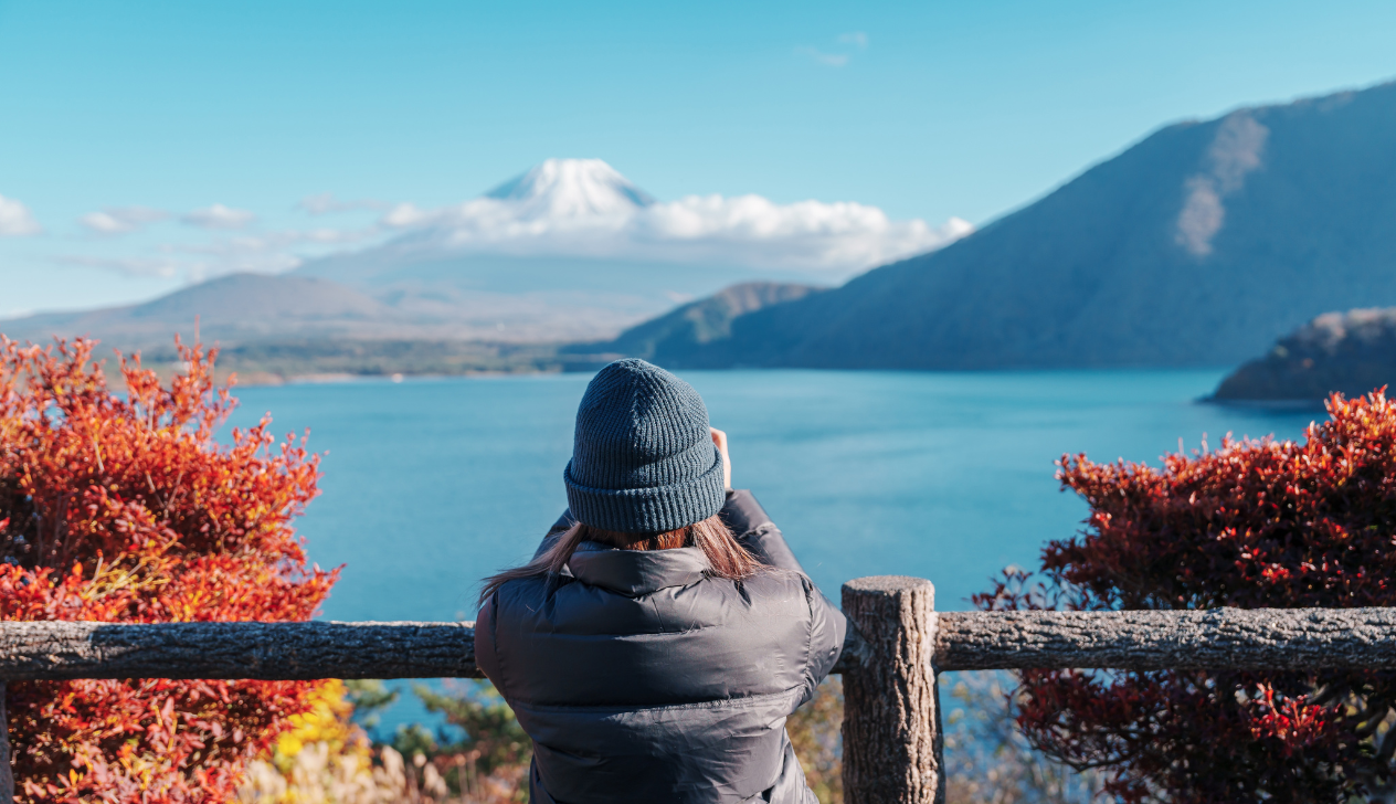 A woman in a blue coat and hat gazing out across a tranquil blue lake at a snow-capped mountain.