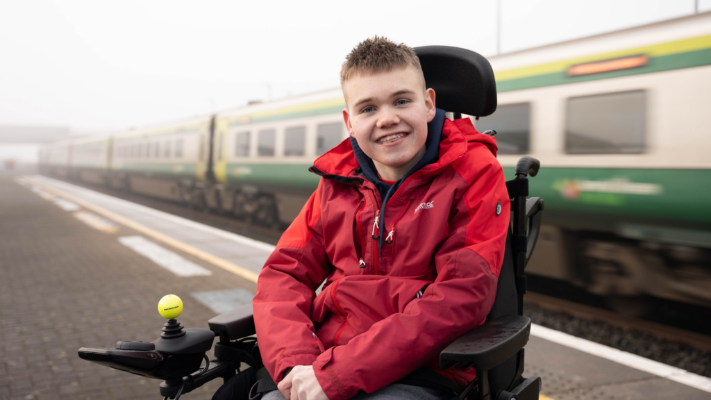 James smiling in his power chair on a train platform with a train whizzing past in the background. James is wearing a waterproof red jacket.