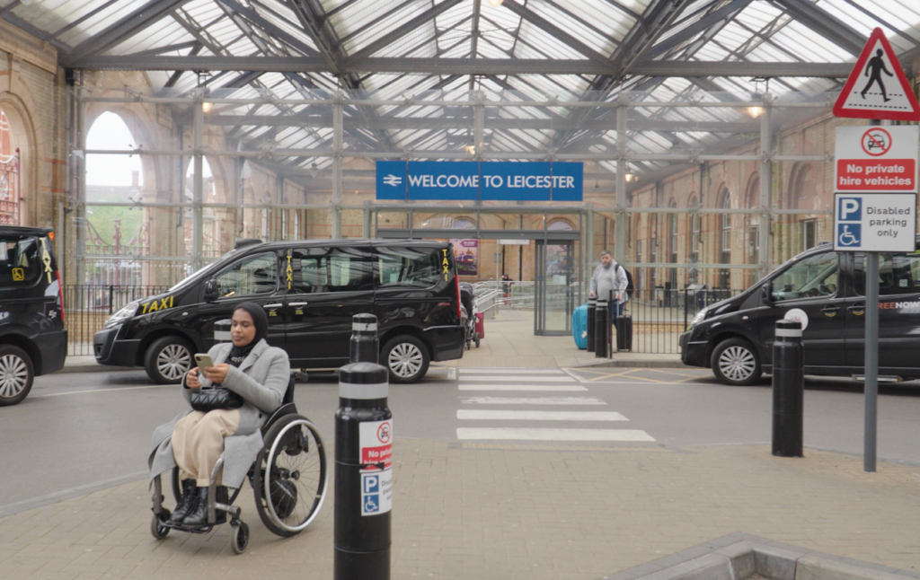 Umaymah, a Disabled wheelchair user, using her phone outside Leicester station before travelling by train with Passenger Assistance.