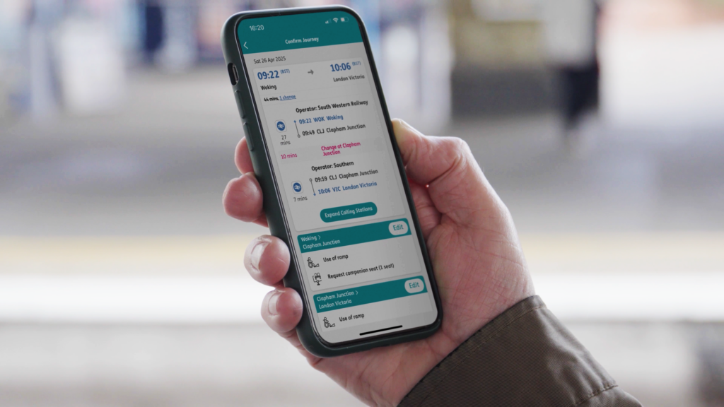 A hand holding a phone displaying the ‘Confirm Journey’ page in the Passenger Assistance app while standing on a train platform.